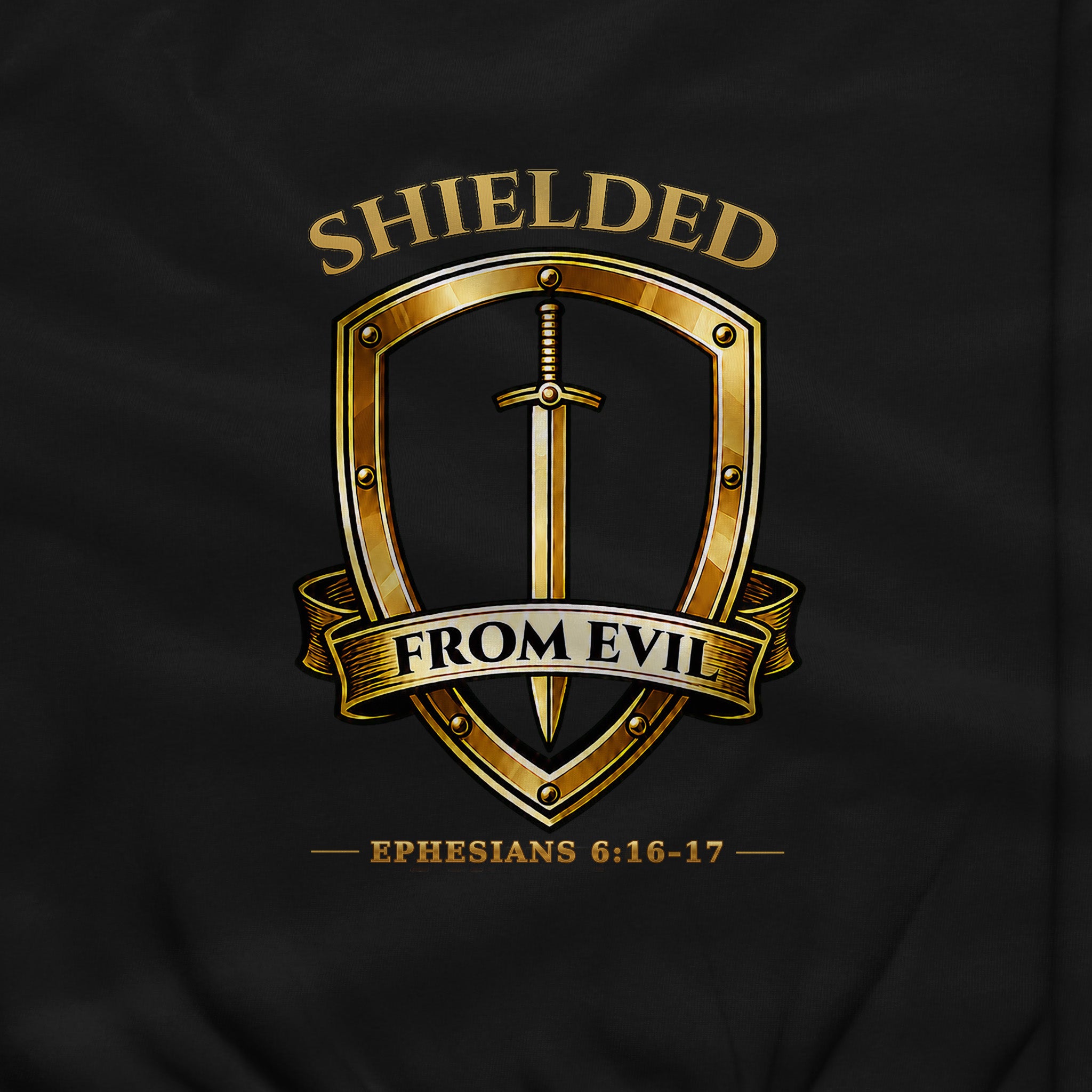 A close-up of a black garment featuring a metallic gold graphic print. The design shows a shield with a vertical sword, a banner reading 'FROM EVIL', the word 'SHIELDED' above, and the Bible verse 'EPHESIANS 6:16-17' below.