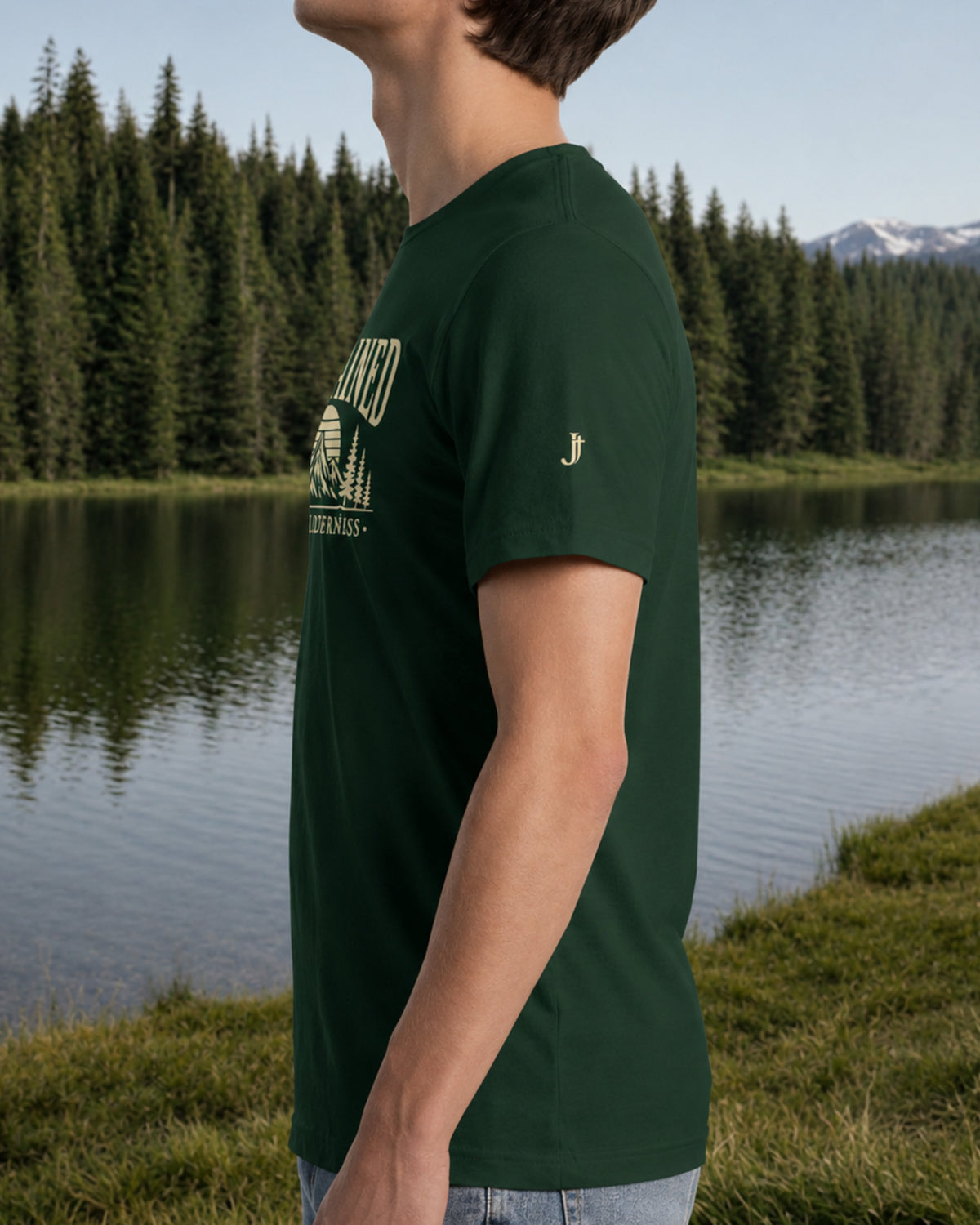 A man wearing a dark green t-shirt with a light green graphic print, standing outdoors next to a calm lake with pine trees and mountains in the background.
