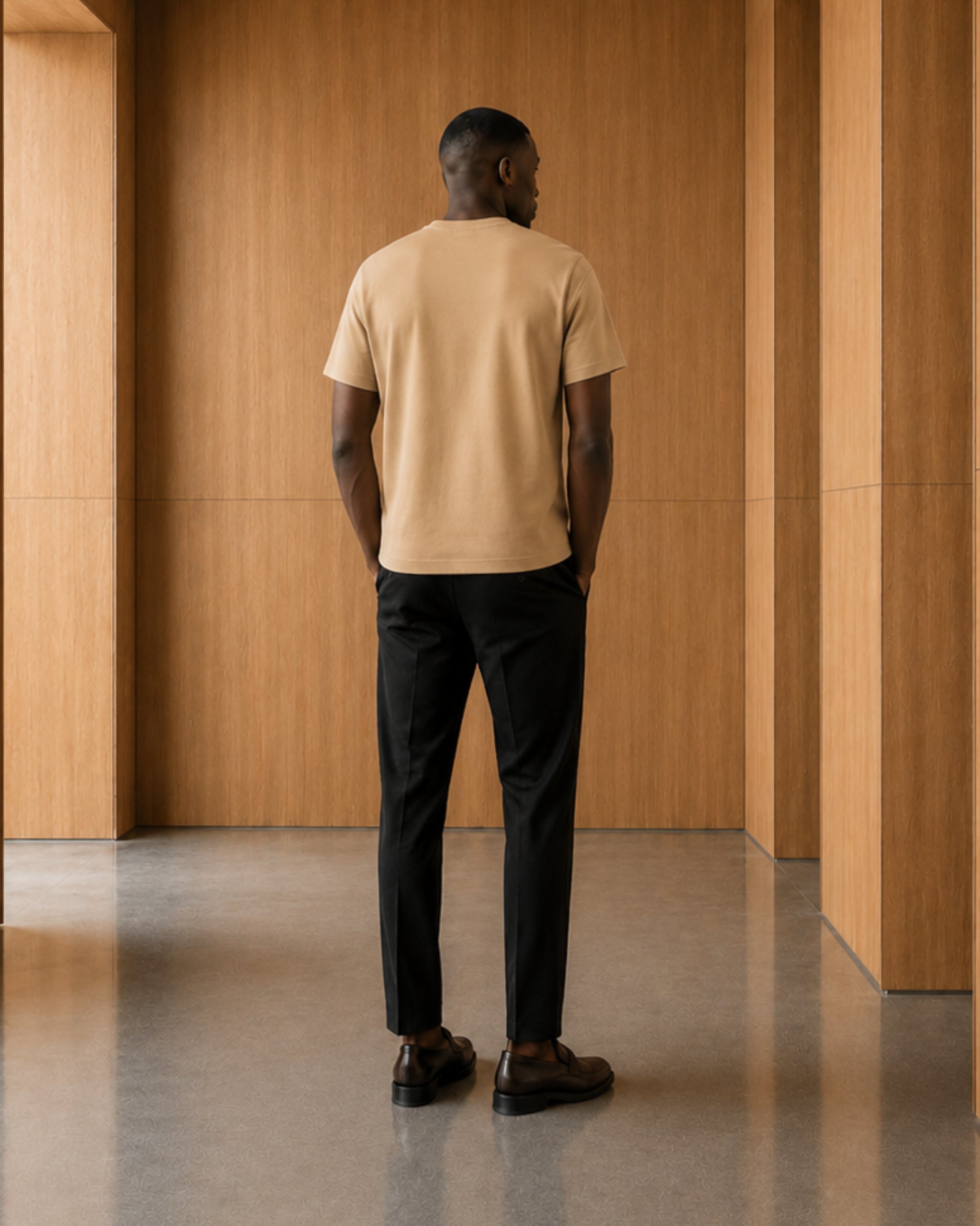 Man wearing a beige t-shirt and black pants standing in a room with wooden walls.