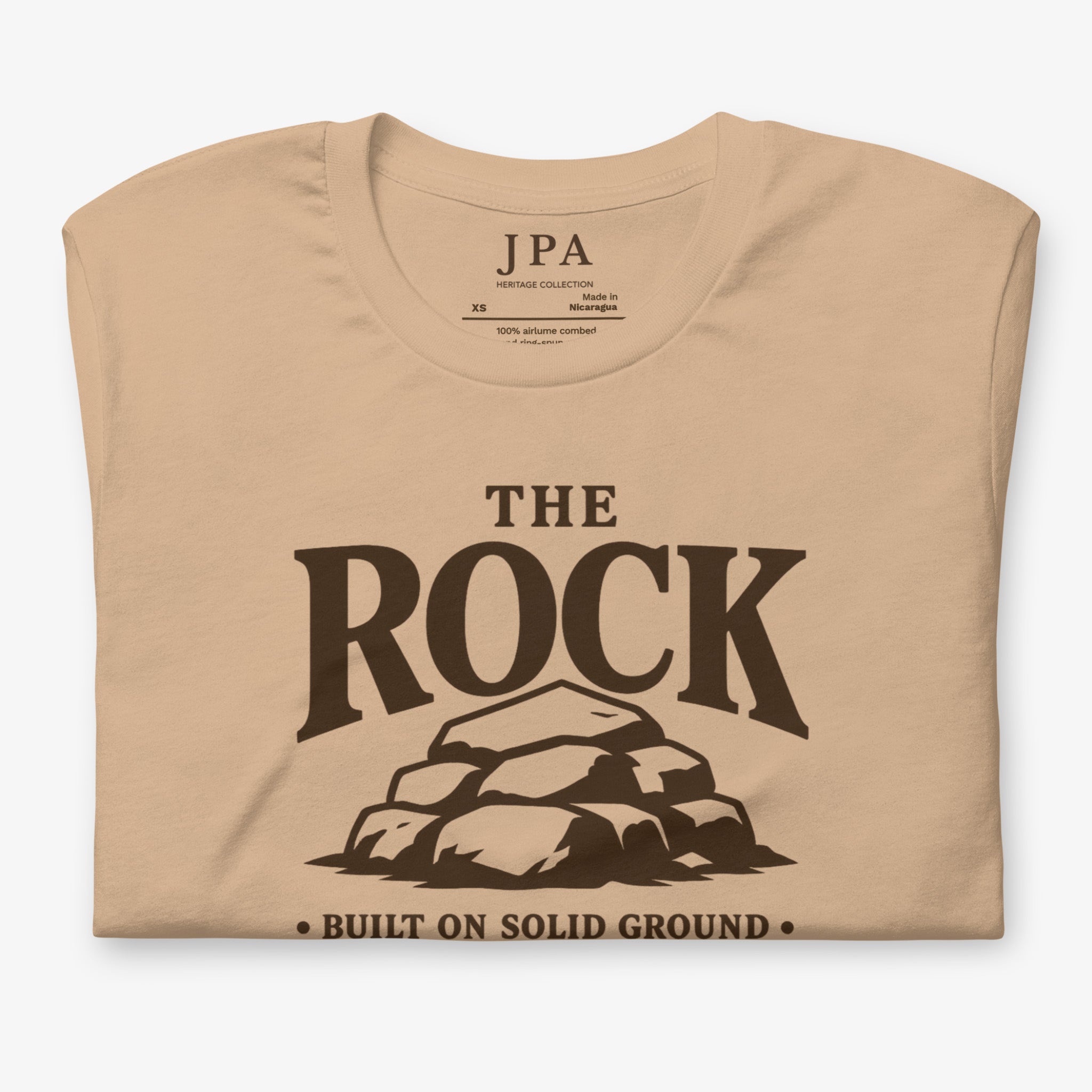 A beige short-sleeve t-shirt with a dark brown graphic print reading 'THE ROCK' above a pile of rocks and 'BUILT ON SOLID GROUND' below, shown on a light gray background.