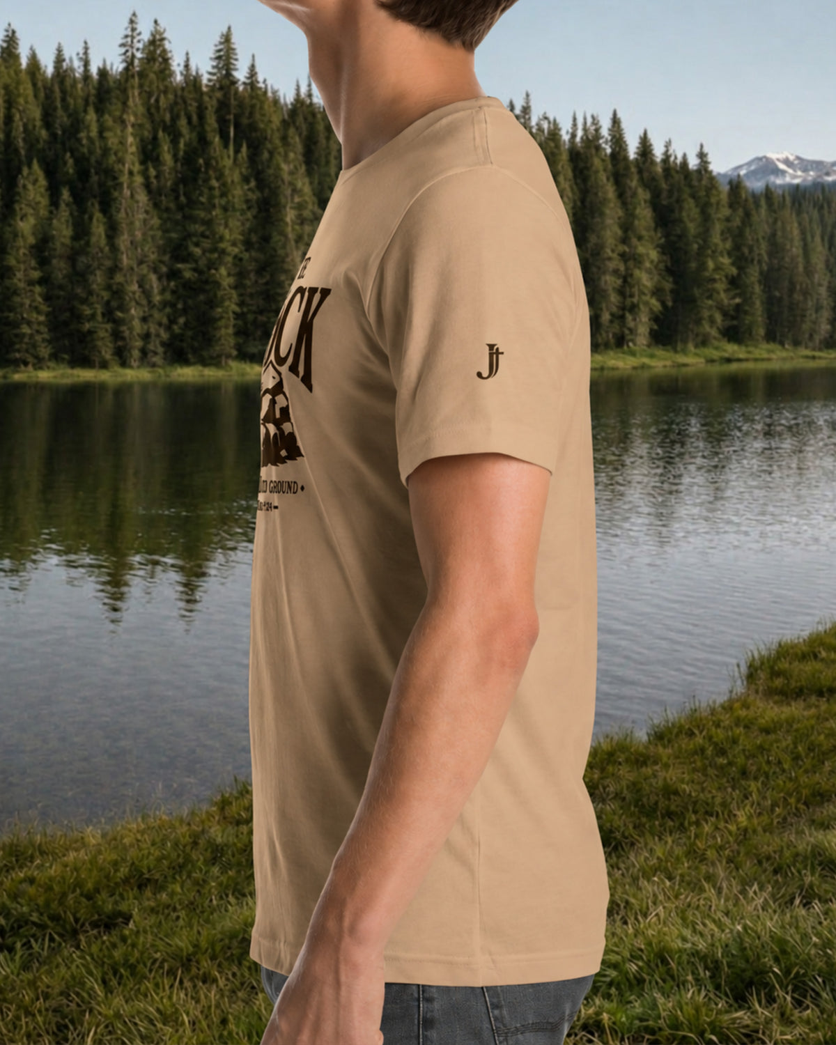 A man wearing a tan crewneck t-shirt with a black graphic print on the front and a small embroidered logo on the sleeve, standing outdoors in front of a lake and pine forest.