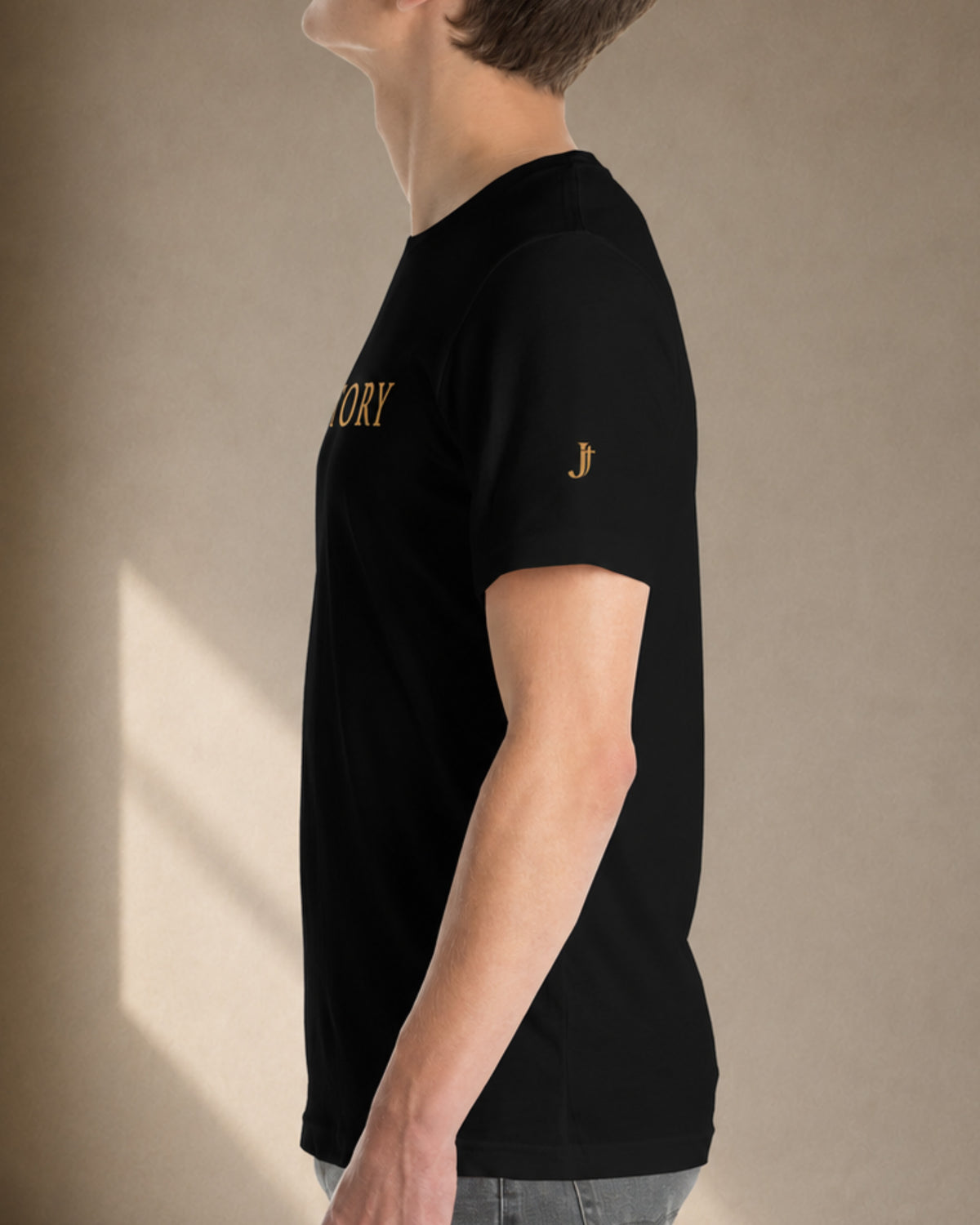 A side-profile view of a person wearing a black short-sleeve t-shirt with gold embroidered text on the chest and a small gold 'JT' logo on the sleeve, set against a plain light gray background.