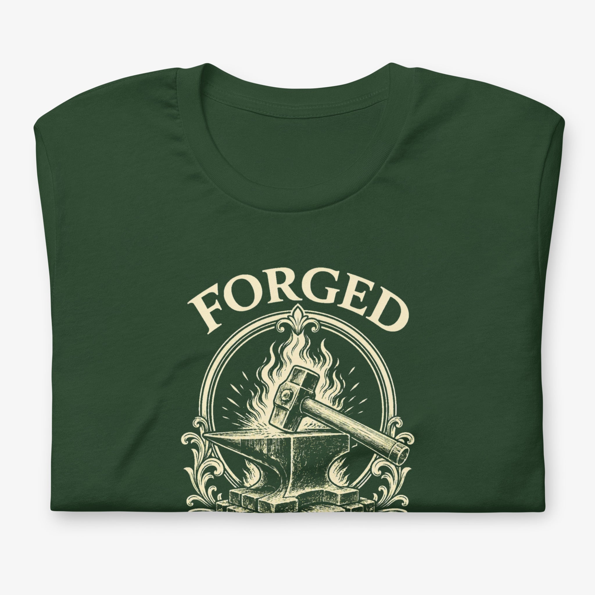 A dark forest green folded t-shirt with a cream-colored graphic on the front. The graphic features the word 'FORGED' in an arched serif font above an illustration of a hammer resting on an anvil with flames.