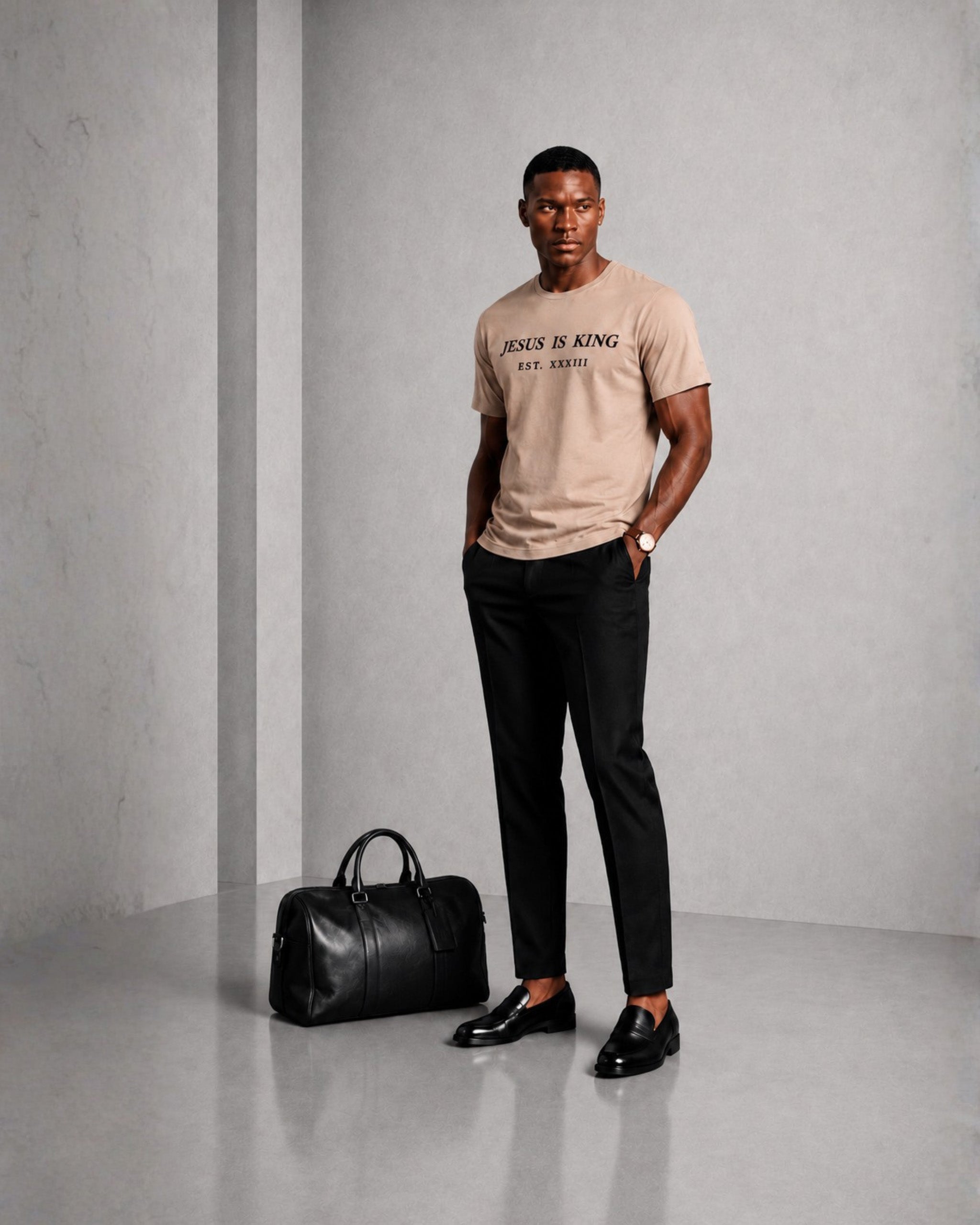 Man wearing a beige t-shirt with text, black pants, and shoes, standing next to a black duffel bag against a gray background.