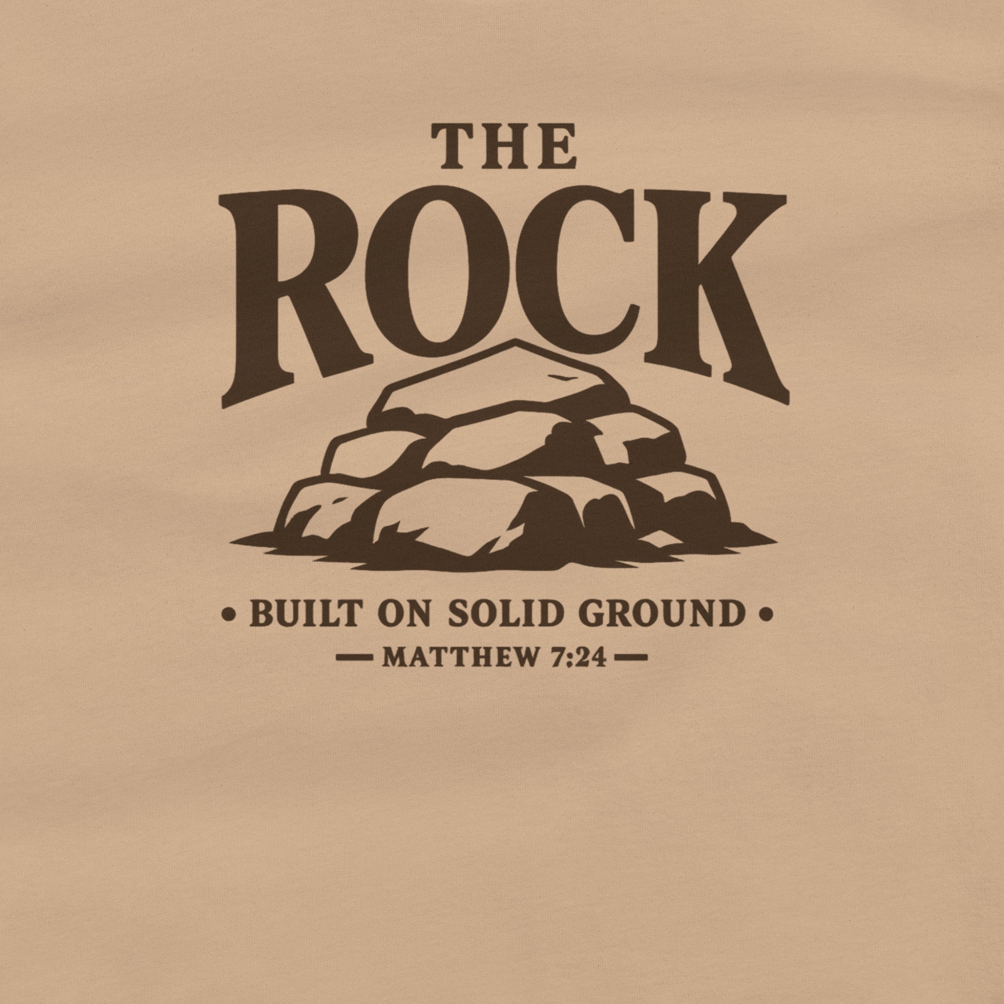 A close-up of a tan or beige fabric surface with a dark brown printed graphic and text reading “THE ROCK,” a pile of rocks illustration, and “BUILT ON SOLID GROUND — MATTHEW 7:24.”.
