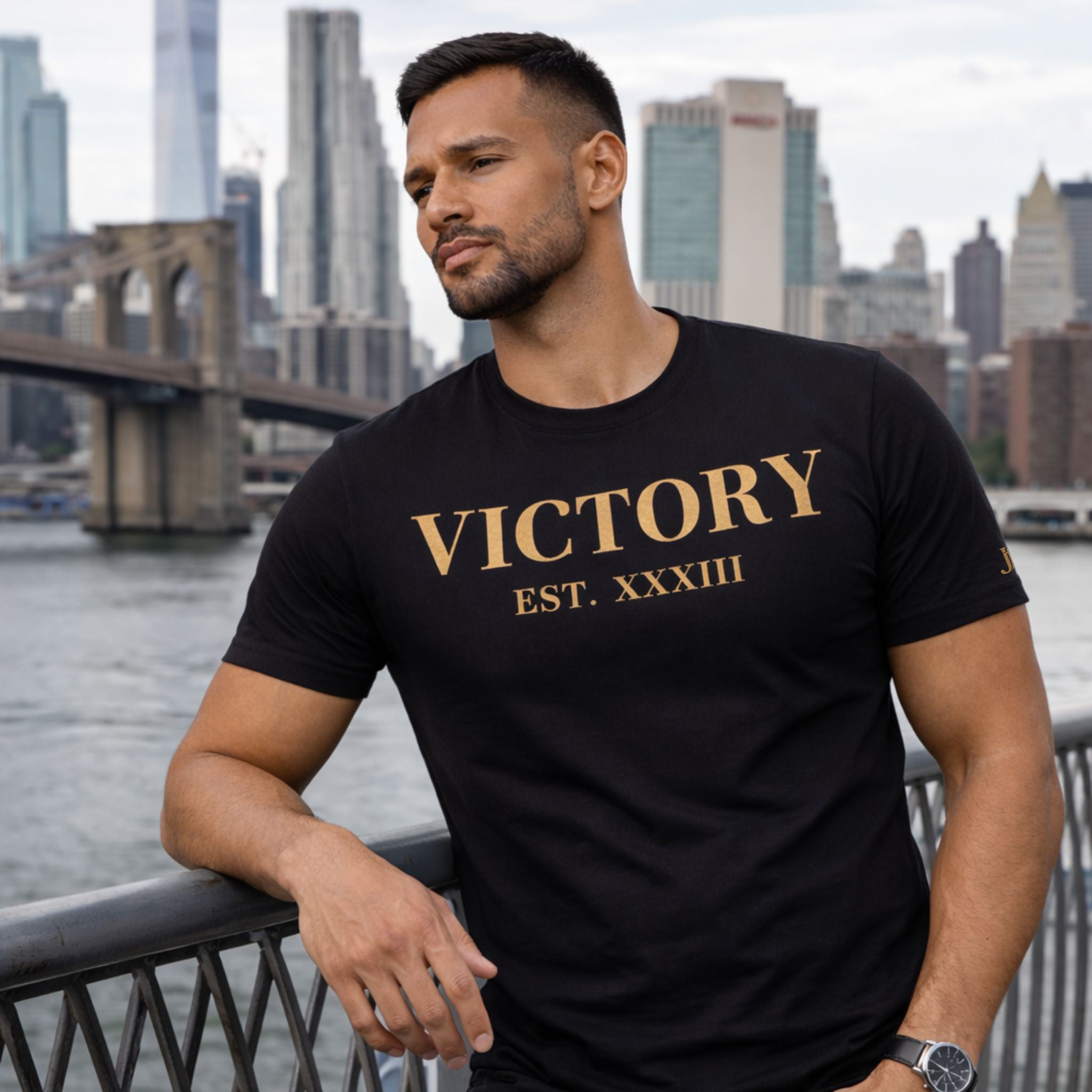 Man wearing a black t-shirt with 'VICTORY EST. XXXIII' in gold, standing in front of a city skyline.